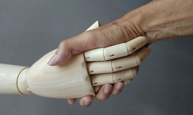 Händeschütteln vor dunkelgrauem Hintergrund, eine Hand ist aus hellem Holz, die andere ist eine menschliche Hand mit dunklerer Hautfarbe